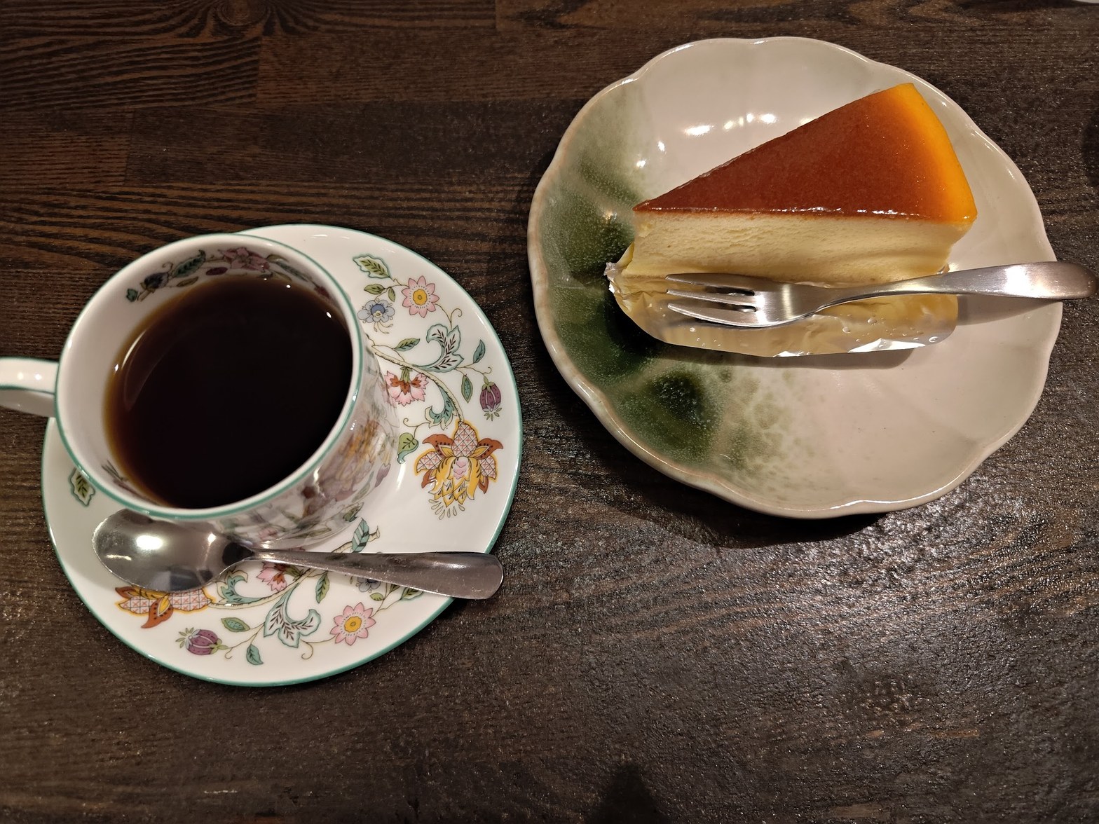 花柄陶器カップに注がれたブラックコーヒーと、三角形のチーズケーキが緑釉の皿に置かれたテーブル